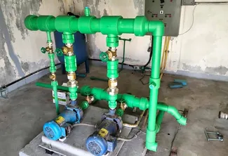 Green PVC water pump and pipe system installed on a concrete floor, featuring two blue electric pumps connected to multiple valves and a control panel mounted on the wall