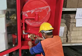 Worker wearing a yellow hard hat and orange safety vest installing or tightening a brass valve inside a red fire hose cabinet containing a coiled hose reel